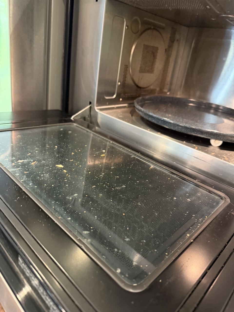 Microwave oven interior before cleaning, with food splatter and crumbs across the open door and turntable